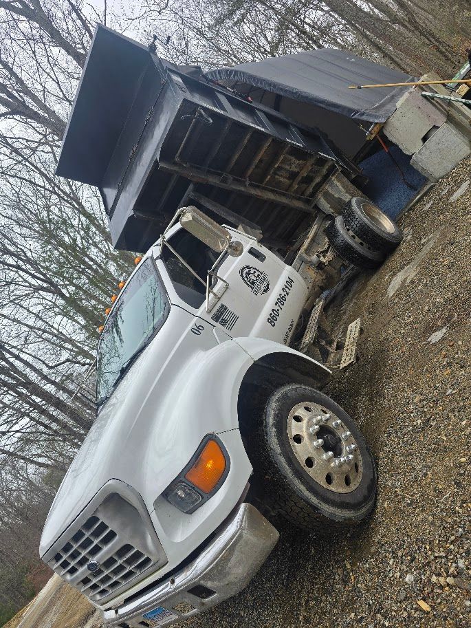 White dump truck with black bed tilted up, parked on gravel next to trees.