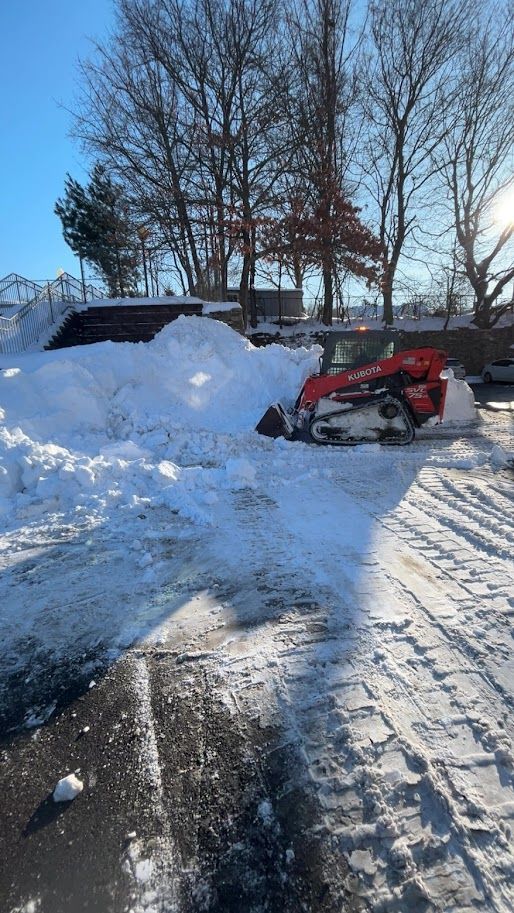 Red skid steer clearing snow from a parking lot on a sunny day.