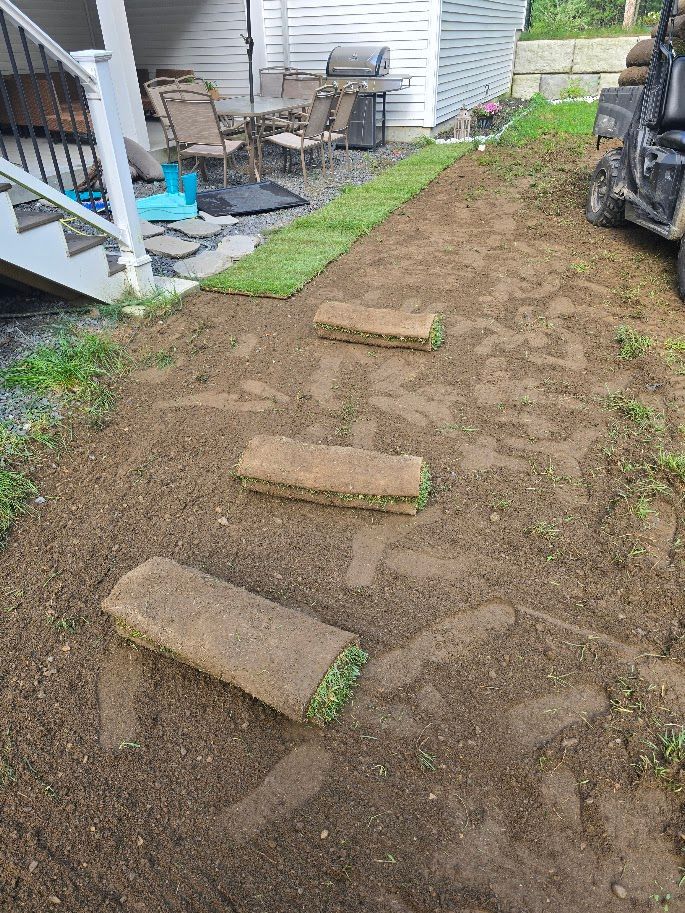 Laying sod rolls on a dirt slope near a house, with steps and a utility vehicle.