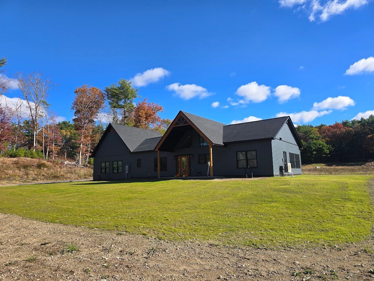 Dark gray house with a black roof, surrounded by green grass and trees under a blue sky.