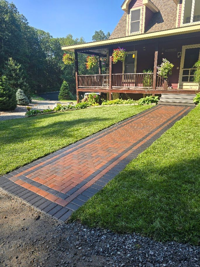 Brick walkway leading to a brown house with a porch. Lush green grass surrounds the path.