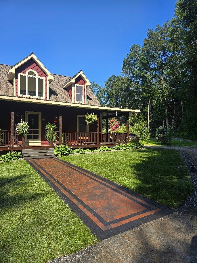 Red-roofed house with covered porch, brick path leading to the entrance, surrounded by green lawn and trees, clear blue sky.