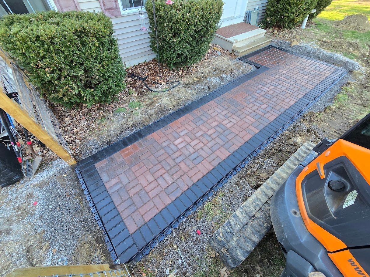 Brick pathway with black border leading to a house entrance, next to bushes and a small tractor.