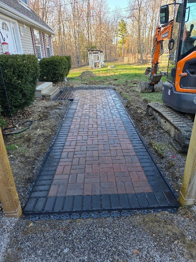 Brick walkway with black border, leading from a house. An excavator is present.