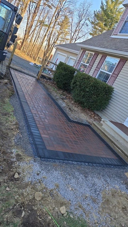 Brick walkway bordering a house with trimmed bushes. Edges lined with black bricks and gravel.