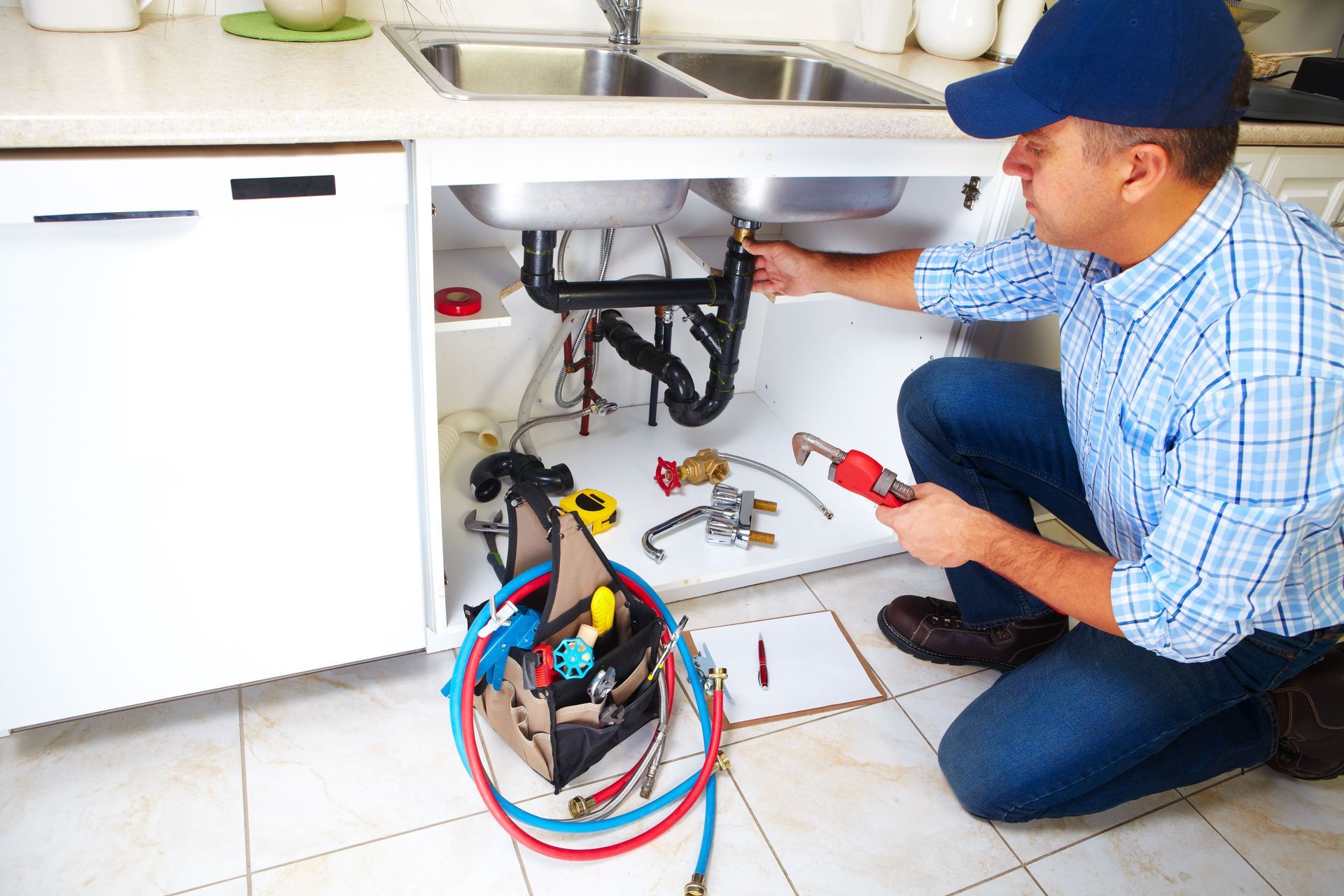 Plumber kneeling under a kitchen sink, fixing pipes. He wears a blue cap and holds a wrench.