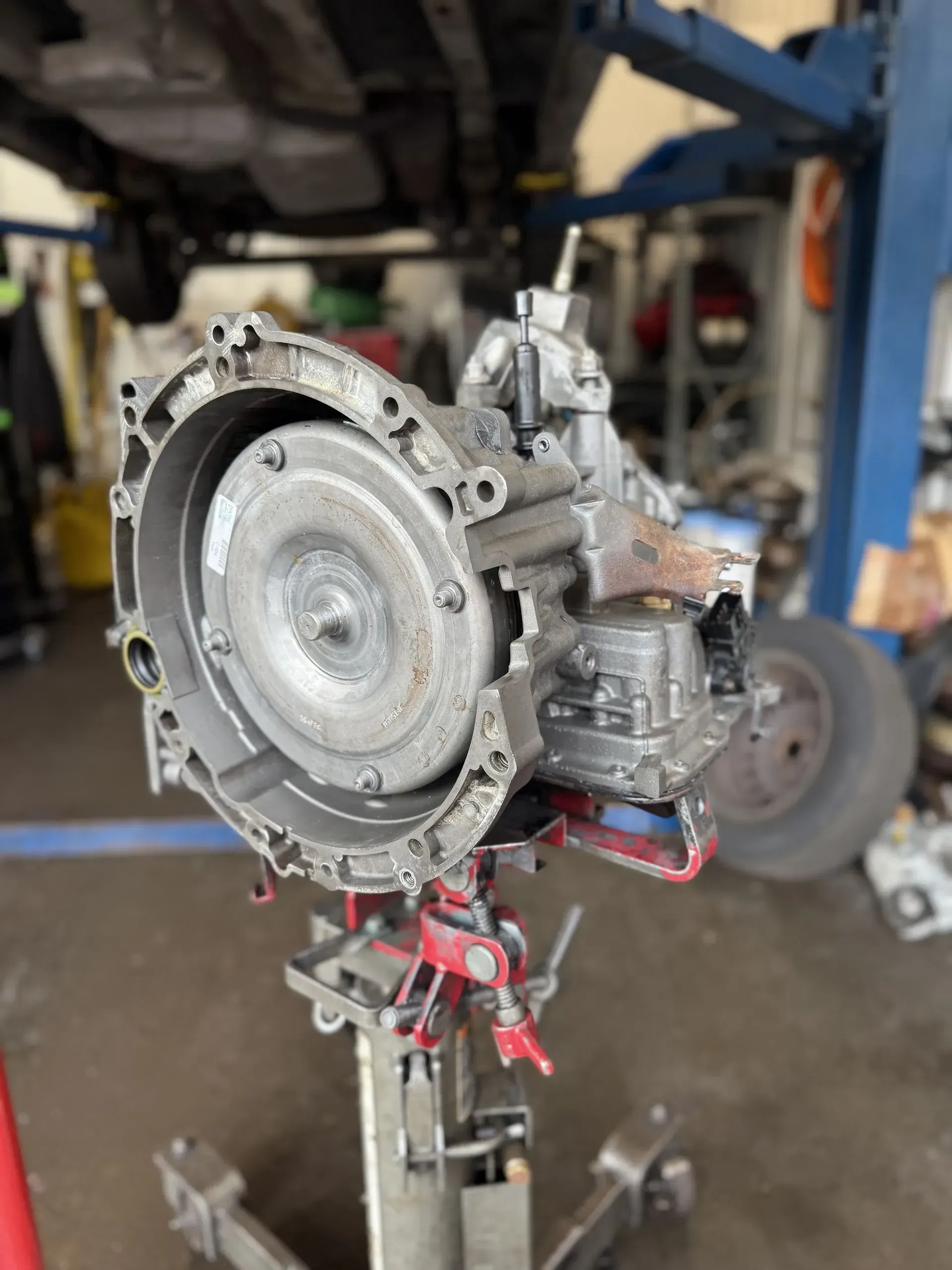 Transmission on a red lift in a repair shop. Gray metal with circular cover. Blue lift visible.