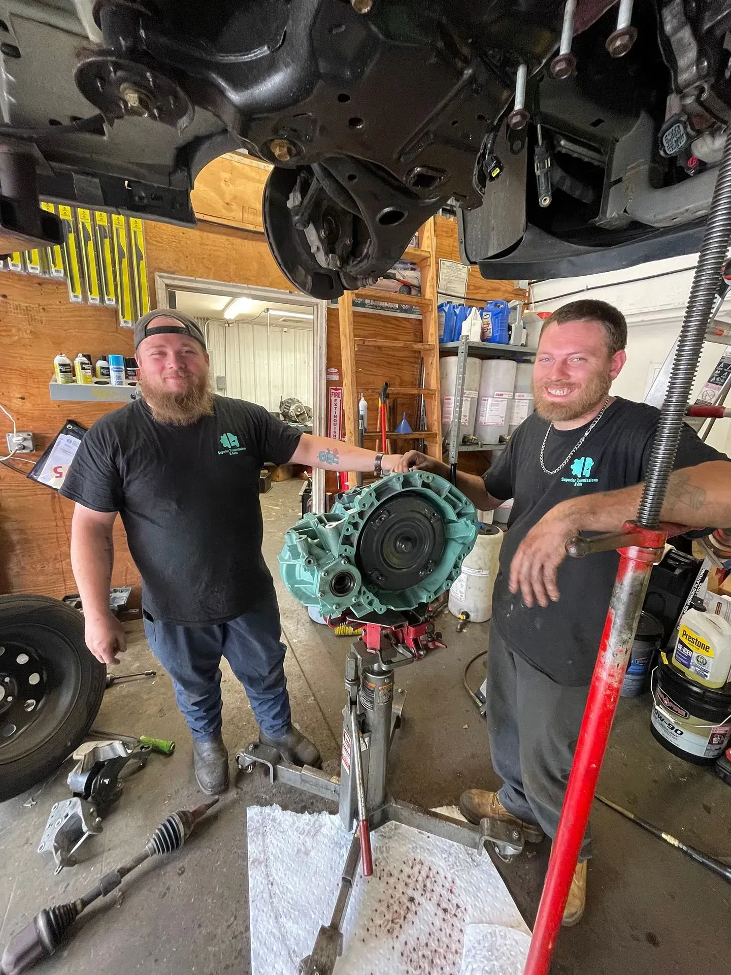 Two mechanics with a teal-colored transmission on a lift under a vehicle in a shop.