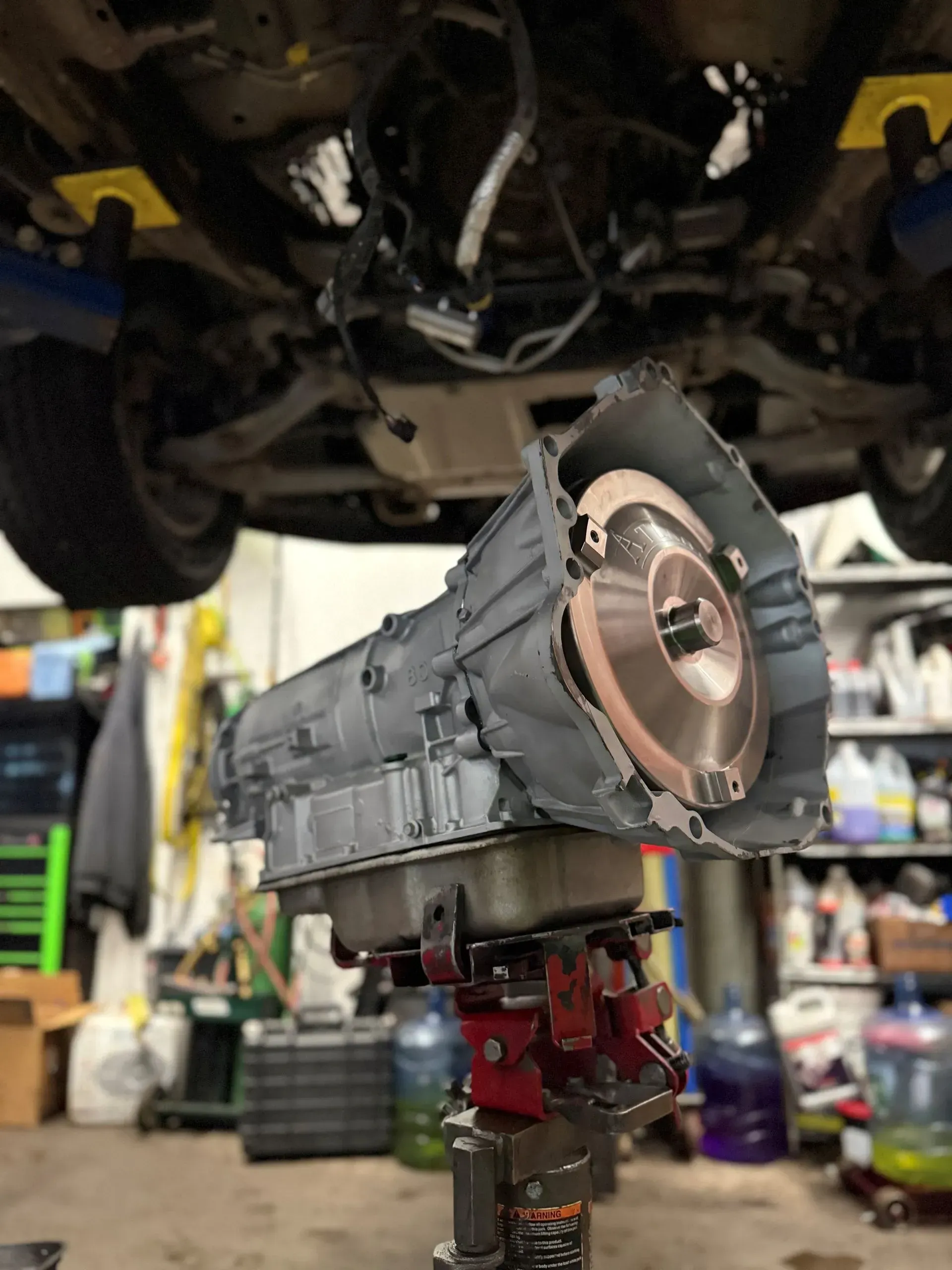 Transmission on a lift, beneath a vehicle. The transmission is grey, and the car is supported by yellow blocks.