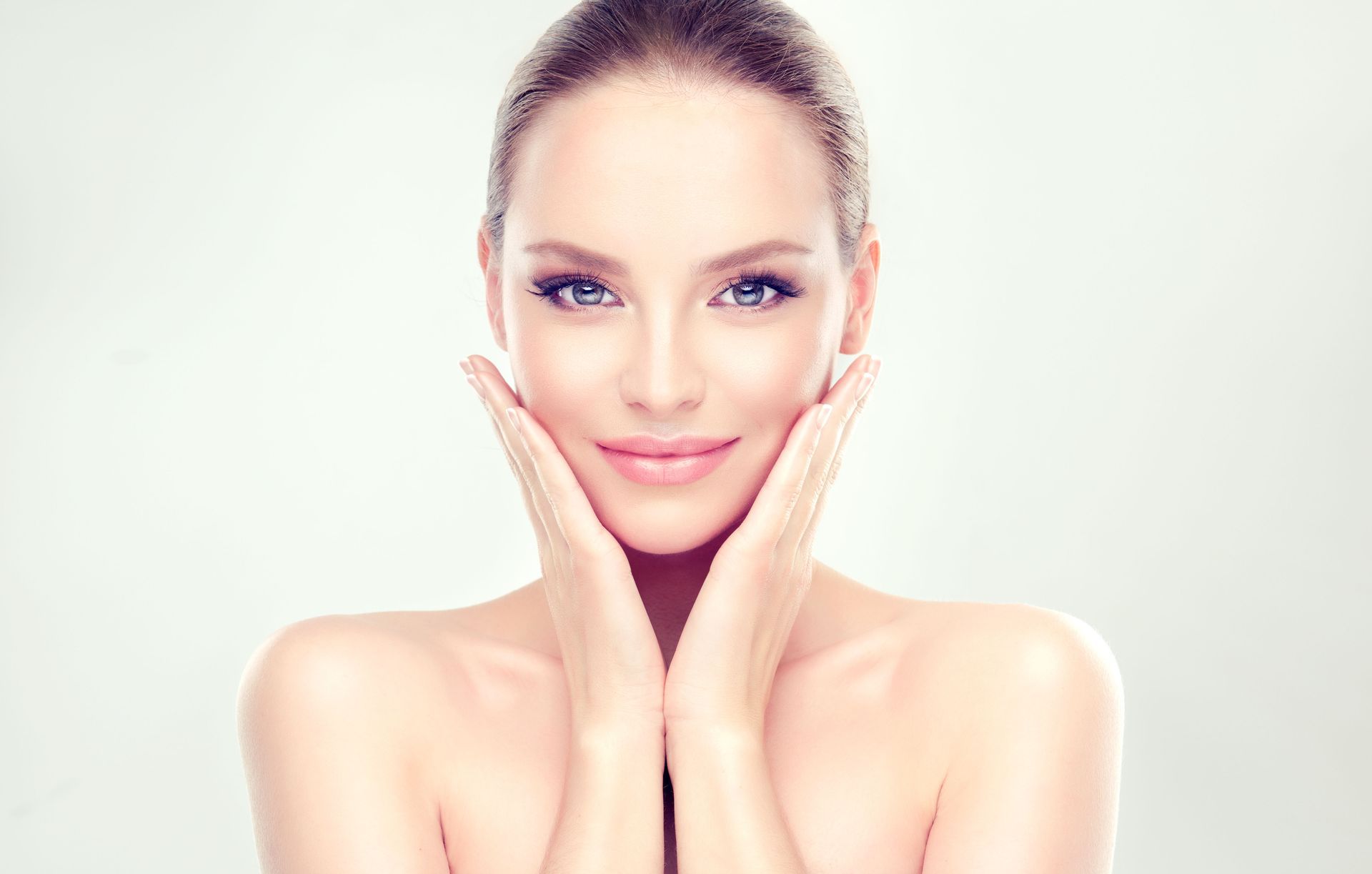 Woman with hands framing her face, smiling, pale skin, light background.