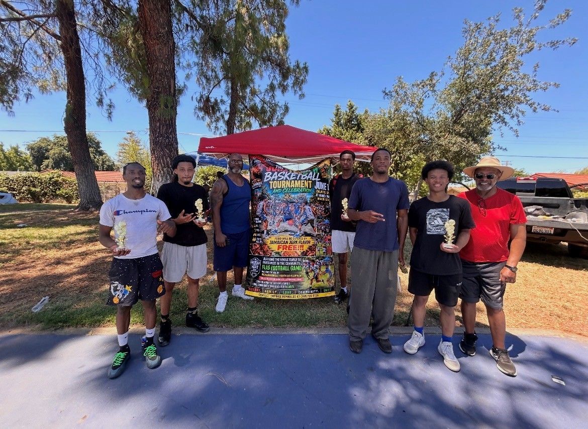 Group of people standing outdoors beside a colorful snack cart under a red canopy in a sunny park.