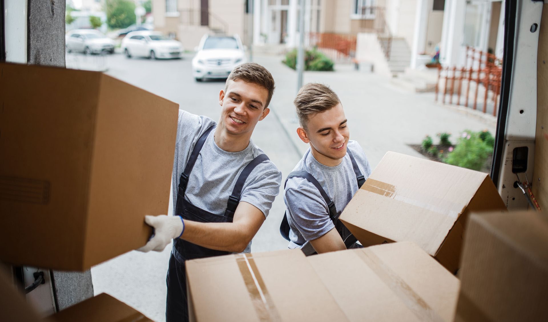 Two young men loading boxes into a truck, smiling. Outdoors, sunny day, wearing gray shirts, overalls.