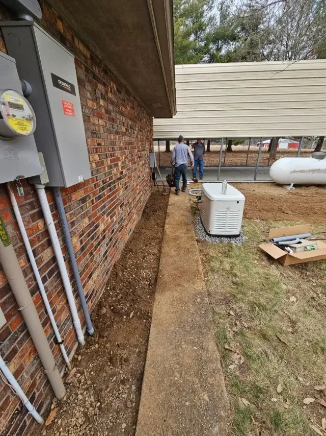 Side yard beside brick house with utility panels, narrow walkway, workers near a covered patio, and yard equipment