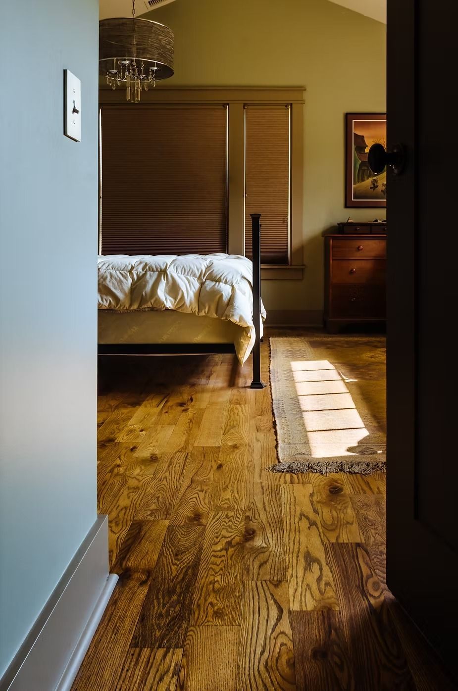 Dark wood floor leads to a bedroom with a bed, dresser, and window with sunlight streaming in.