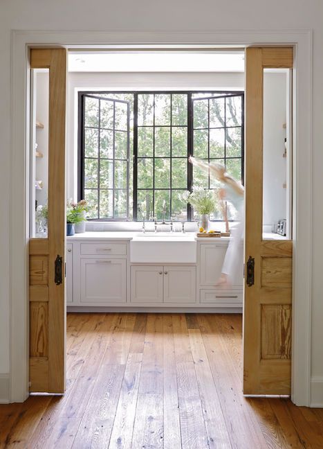 Wooden sliding doors open to a bright white kitchen with a large window, looking out to a green tree.