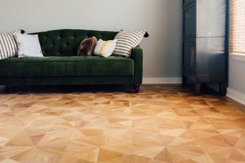 Green velvet sofa with decorative pillows against a light gray wall, hardwood floor with geometric pattern.