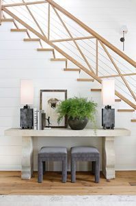 Console table with lamps, artwork, and a plant under a wooden staircase. Gray stools sit in front.