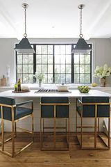 Modern kitchen with white island, blue velvet stools with gold frames, two pendant lights, and a large window.