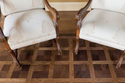 Two antique chairs with white patterned cushions on a dark brown parquet floor.