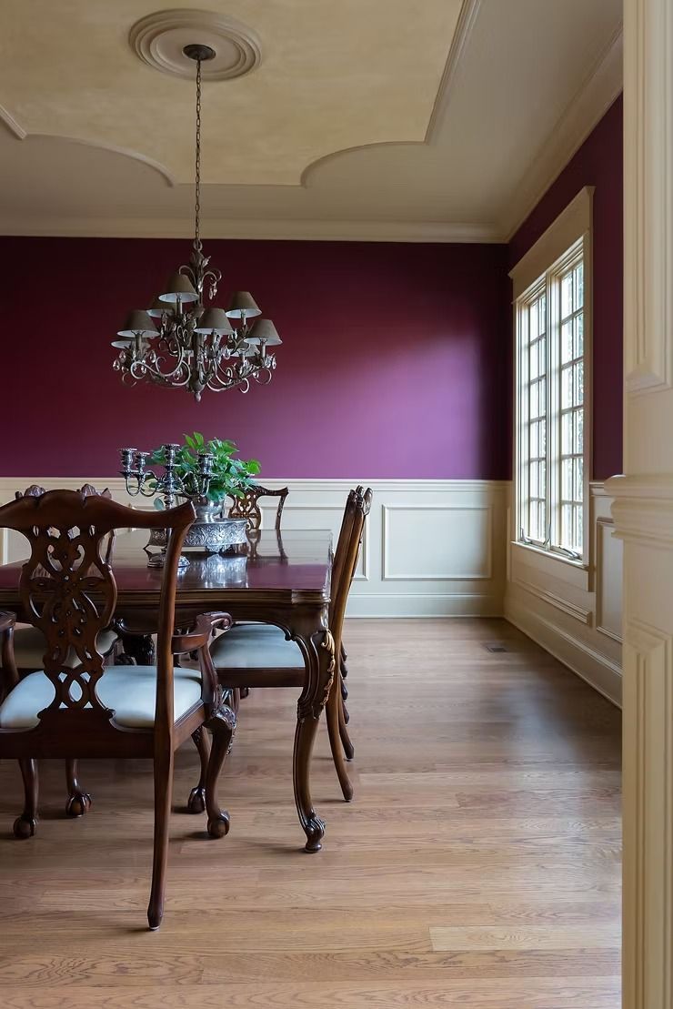 Formal dining room with a burgundy wall, chandelier, wooden table and chairs, and a window.