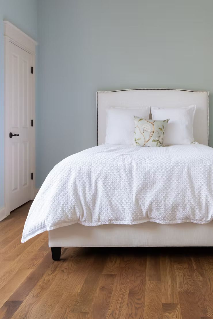 Bedroom with white bed, bedding, and headboard against a pale blue wall. Wooden floor and white door on the left.