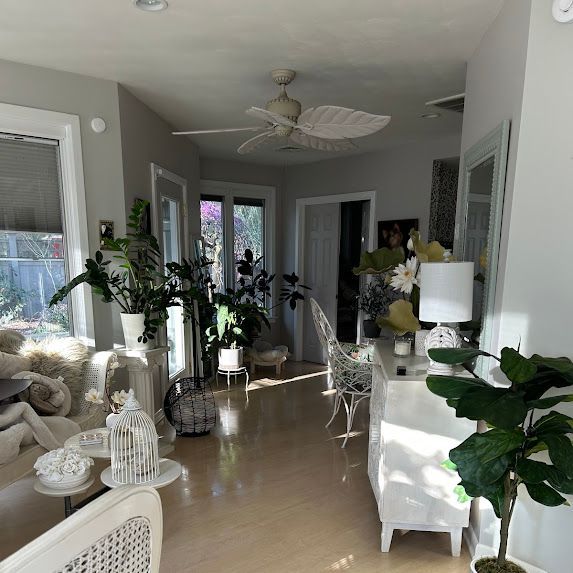 Bright living room with white furniture, large plants, and a ceiling fan in a sunlit hallway area