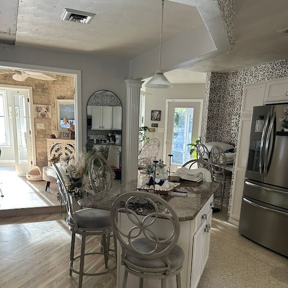 Bright kitchen with stainless fridge, white cabinets, granite island, bar stools, and a doorway to another room.