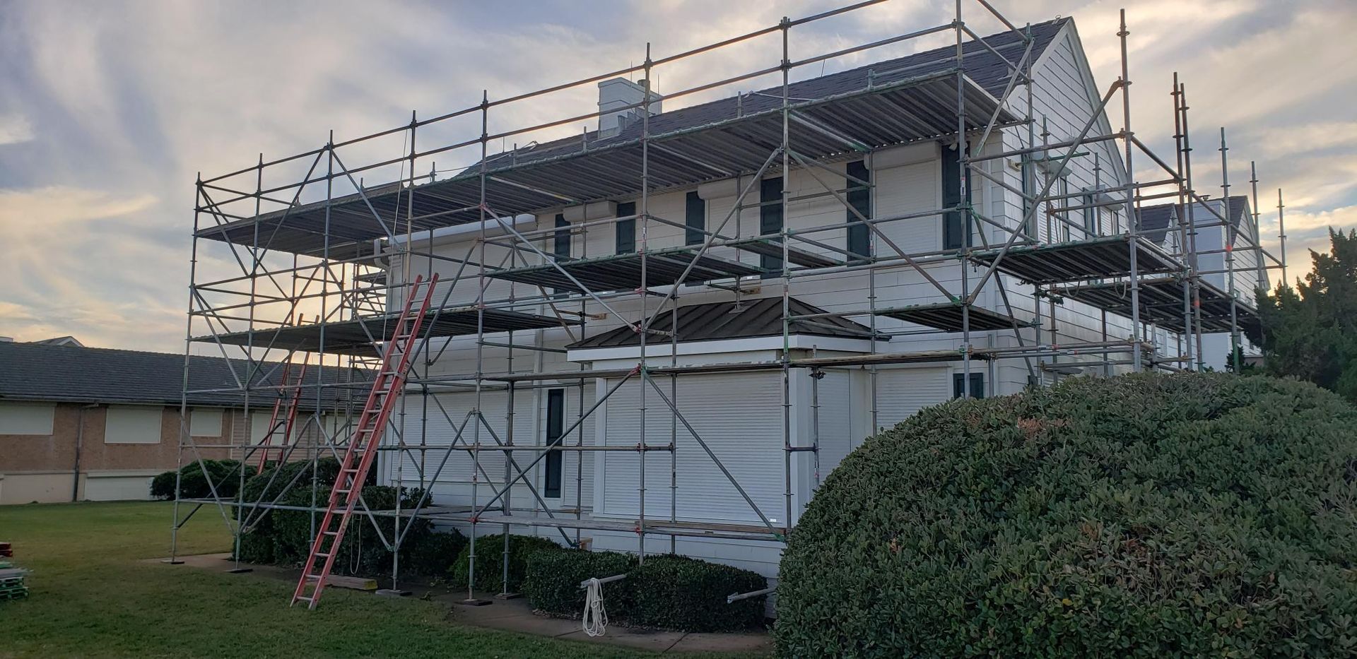 House under construction with scaffolding, white wrap, and a ladder in a suburban yard at sunset