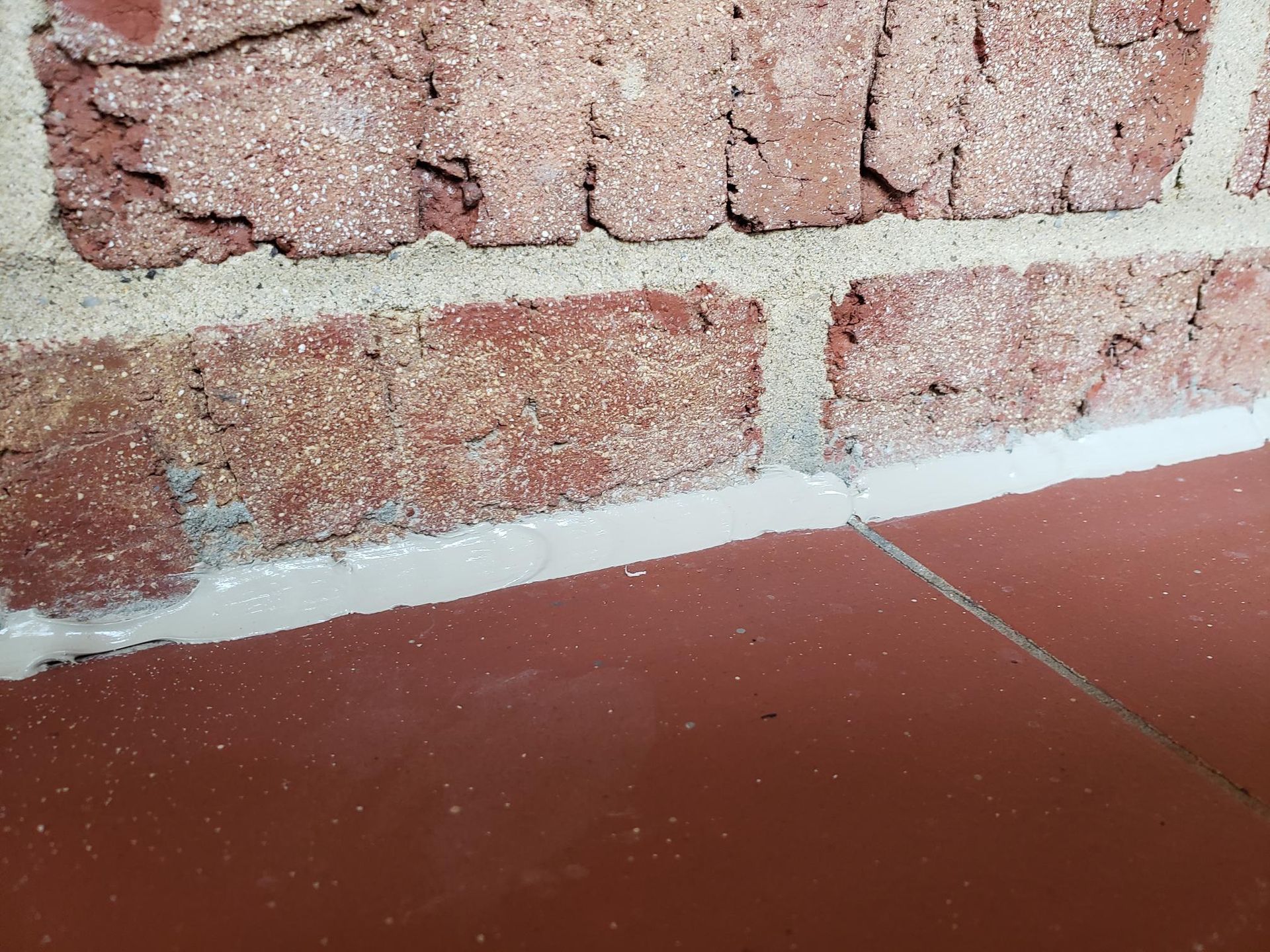 Close-up of a brick wall meeting a reddish tiled floor with white caulk along the base.