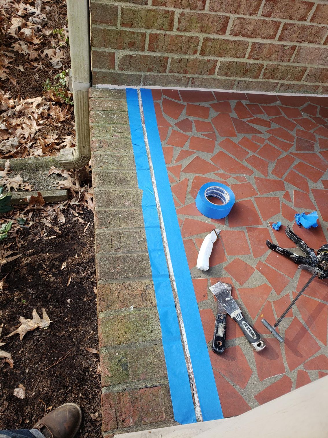 Blue painter’s tape marks a brick porch corner beside tools and a roll of tape.