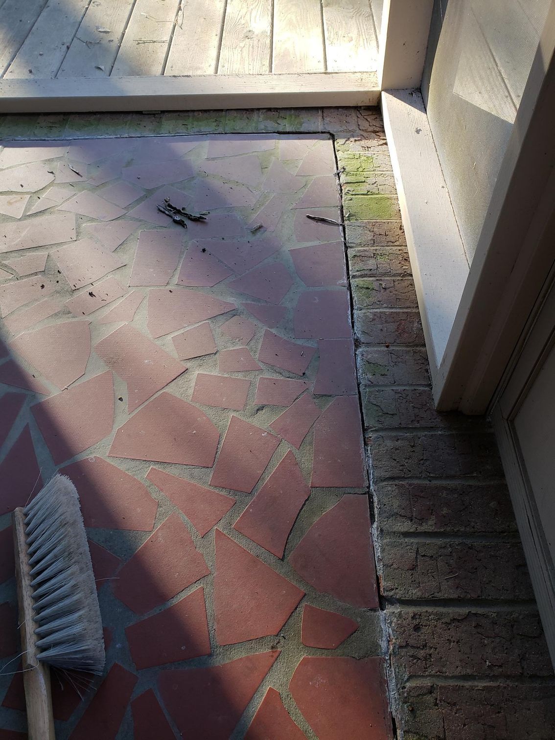 Sunlit front porch with red tiled floor, brick edge, and a broom in the lower left corner