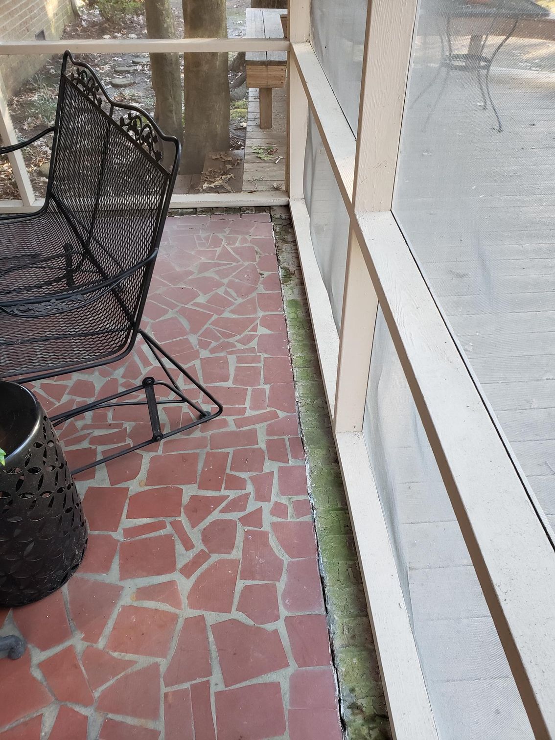 Narrow red-tiled patio beside a house, with a black wire chair and railing.