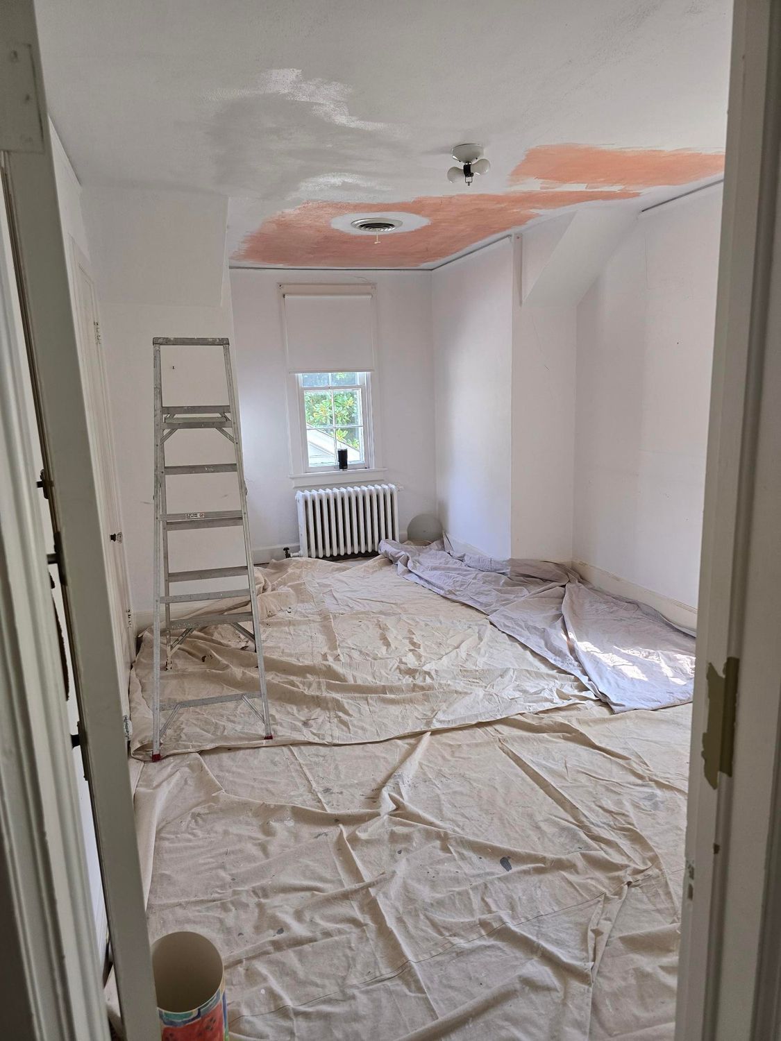 Renovating room with ladder, plastic-covered floor, and unfinished ceiling near a window and radiator