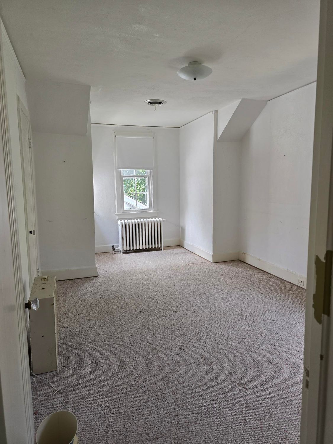 Empty narrow room with white walls, carpeted floor, a window, and a radiator near the far end.