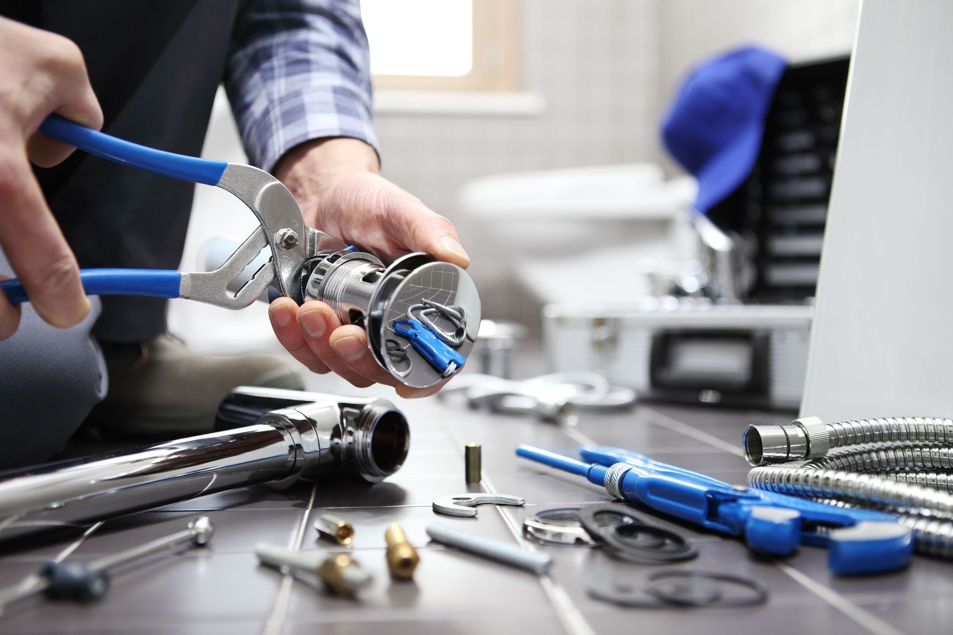 A plumber uses blue-handled pliers to assemble a chrome sink faucet component, with tools and hardware on a tiled floor.