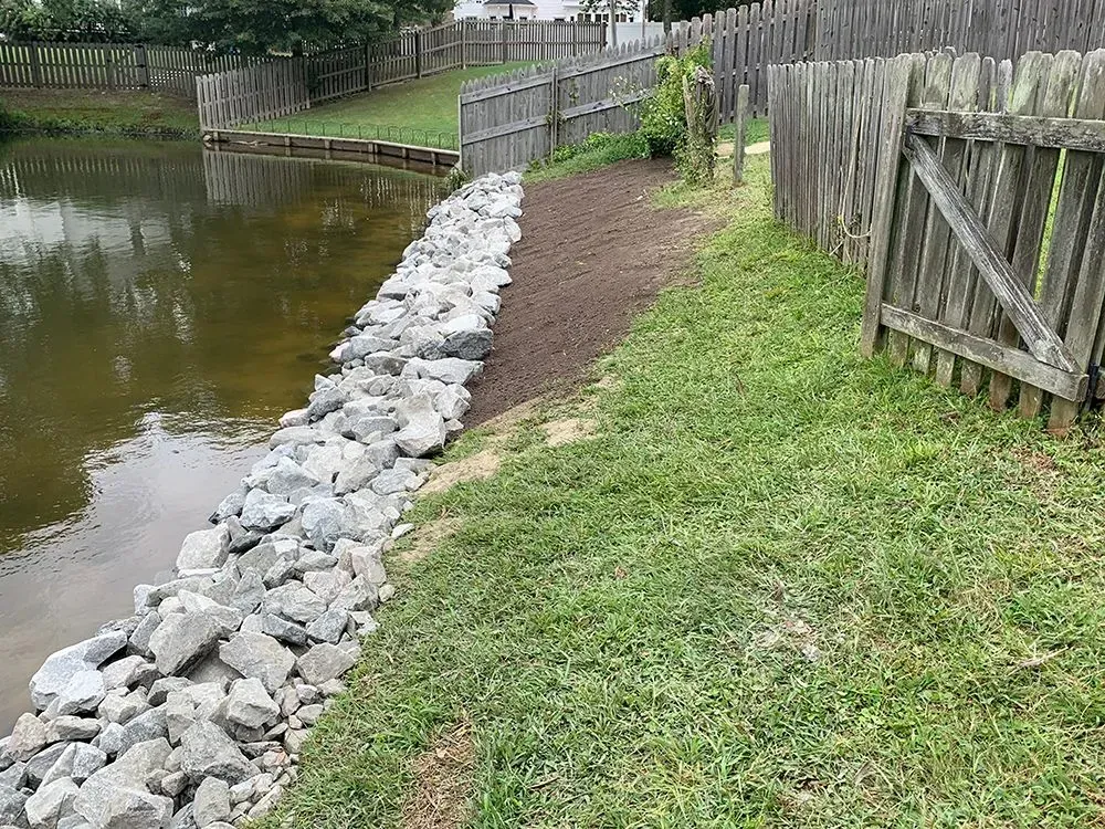 A wooden fence surrounds a rock wall next to a pond.