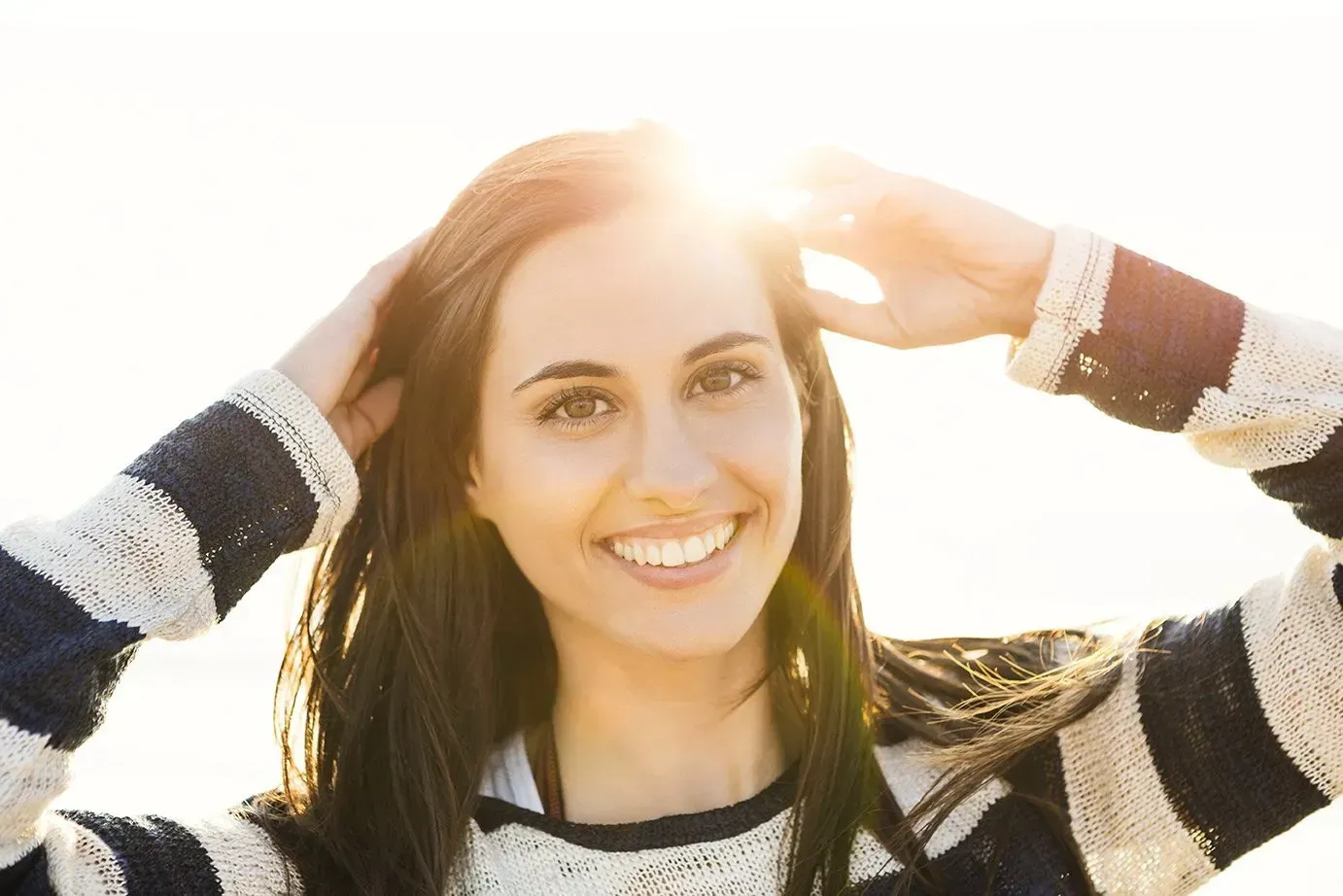Woman smiling, arms raised, sunlit background, wearing striped sweater.