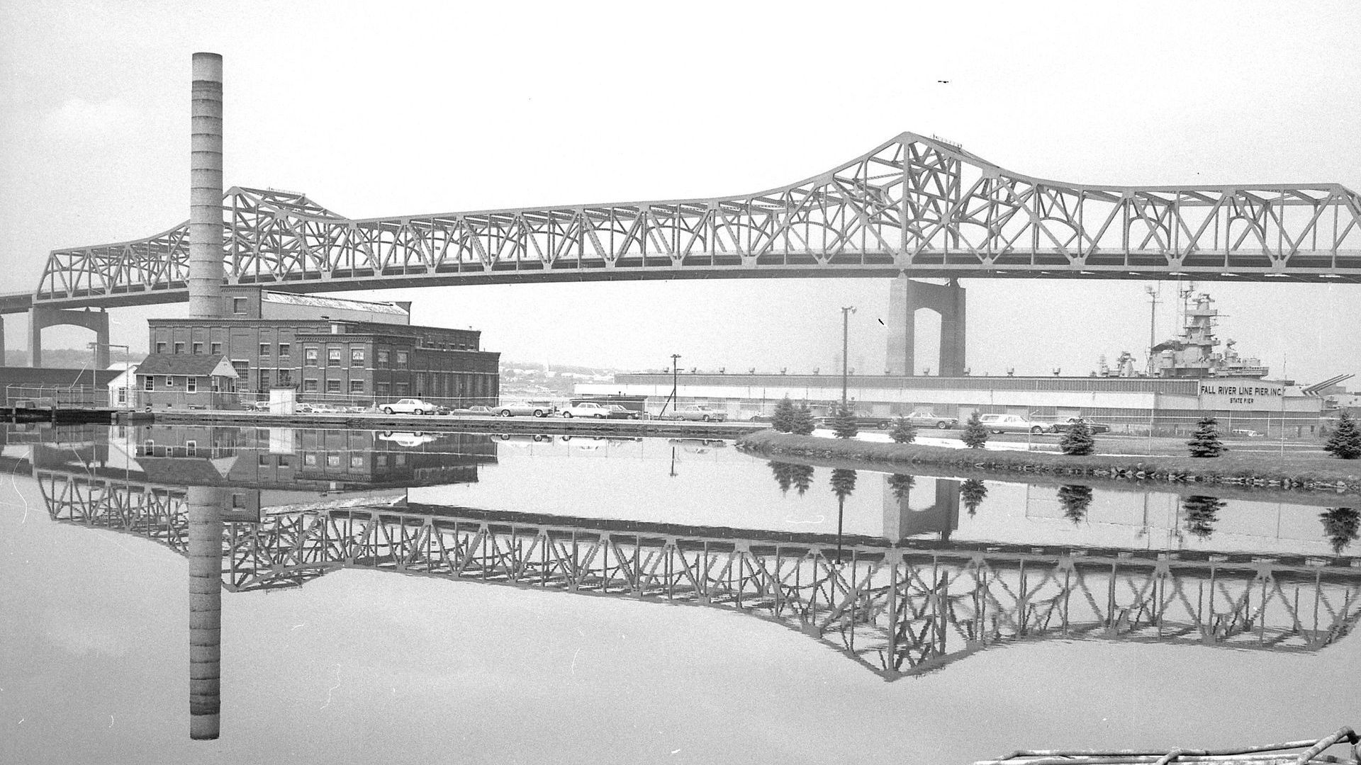 Bridge over water, factory with smokestack, and a ship in a harbor, reflected in the water.