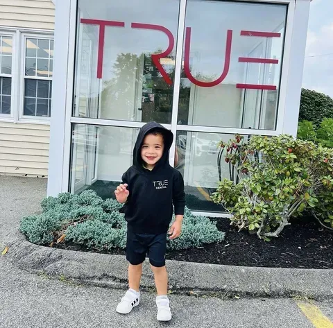 Child in black hoodie and shorts waves in front of a window with the word "TRUE" on it.