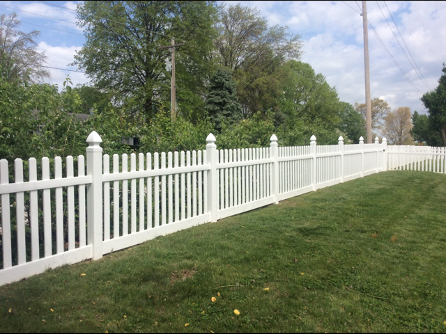 White picket fence along a grassy roadside with trees and utility poles under a cloudy sky