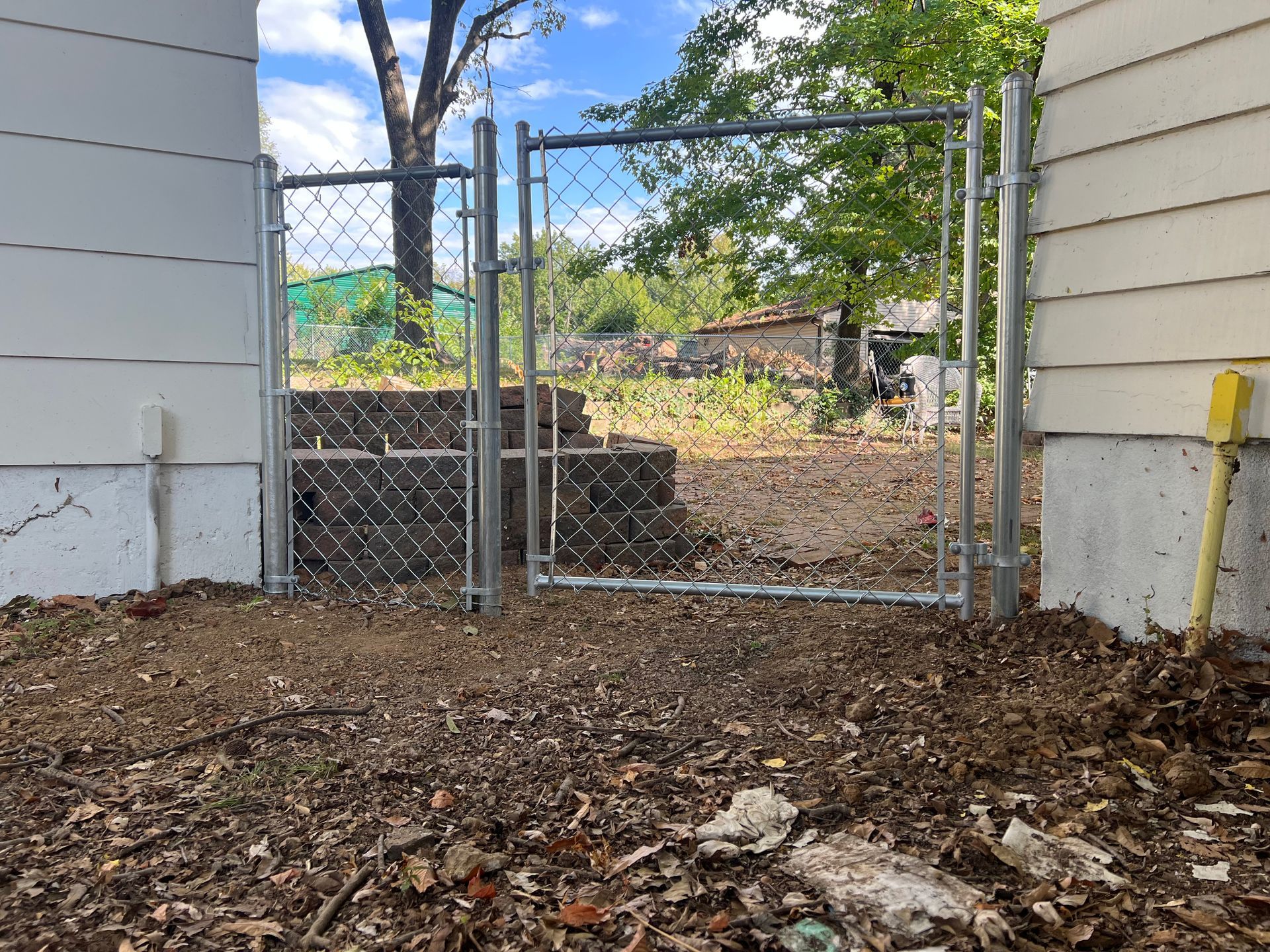 Chain-link gate between two buildings, with dirt ground and trees visible beyond.