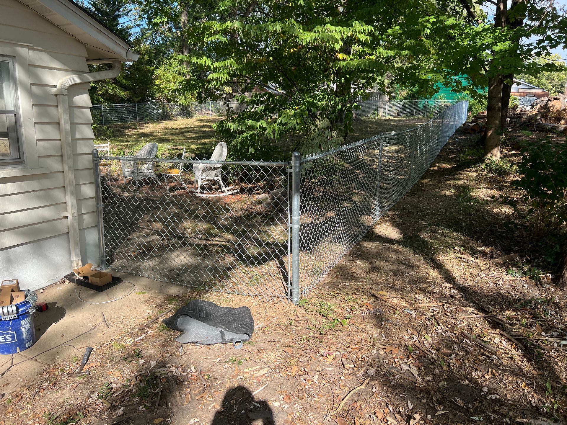 Chain-link fence panel leaning in a wooded backyard beside a house, with a small chair and scattered items nearby