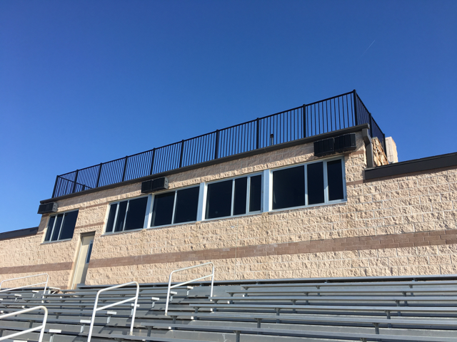 Empty bleachers below a tan stadium wall and rooftop railing under a clear blue sky