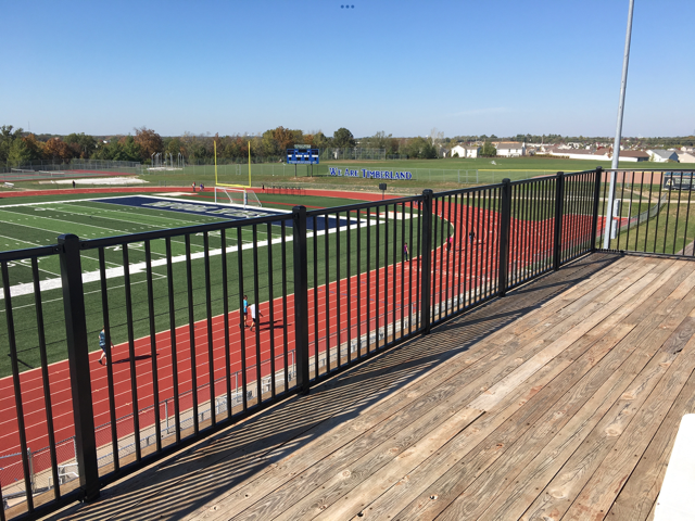 Empty bleachers overlooking a football field with a red track and black railing on a sunny day