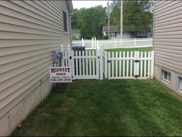 Narrow grassy side yard between houses with a white picket gate and a small sign on the left wall