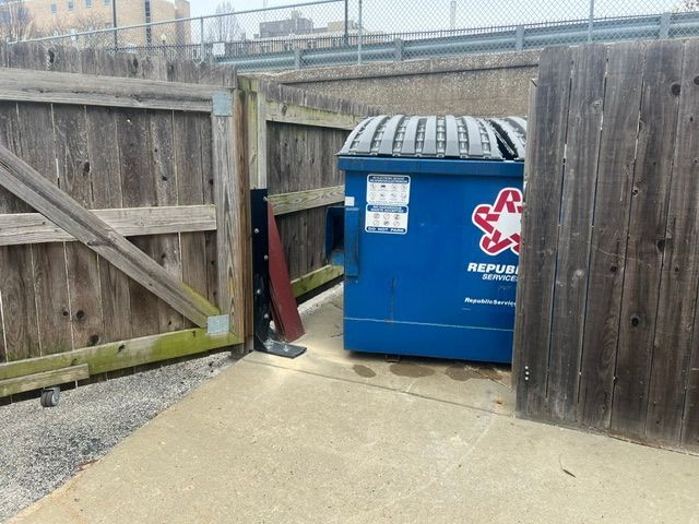 Blue dumpster in a narrow fenced alley with an open wooden gate