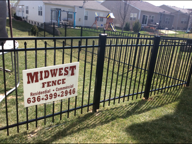 Black metal fence with a sign reading “MIDWEST FENCE” in a grassy residential yard.