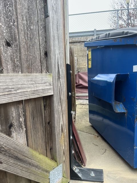 Narrow alley between wooden fence and blue dumpster with a metal ramp on concrete ground