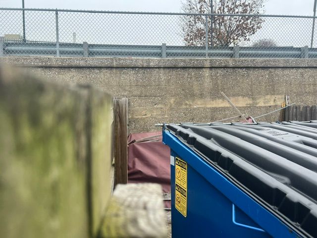 Blue dumpster beside a chain-link fence, with a maroon tarp and concrete wall in the background.