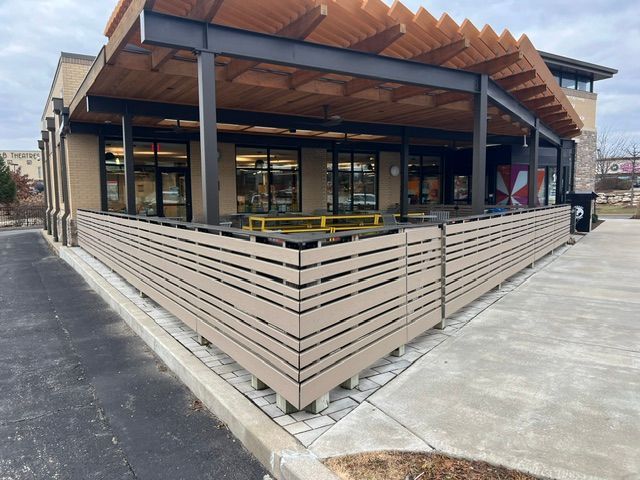 Modern storefront with wooden awning, glass windows, and fenced outdoor seating area on a concrete corner.